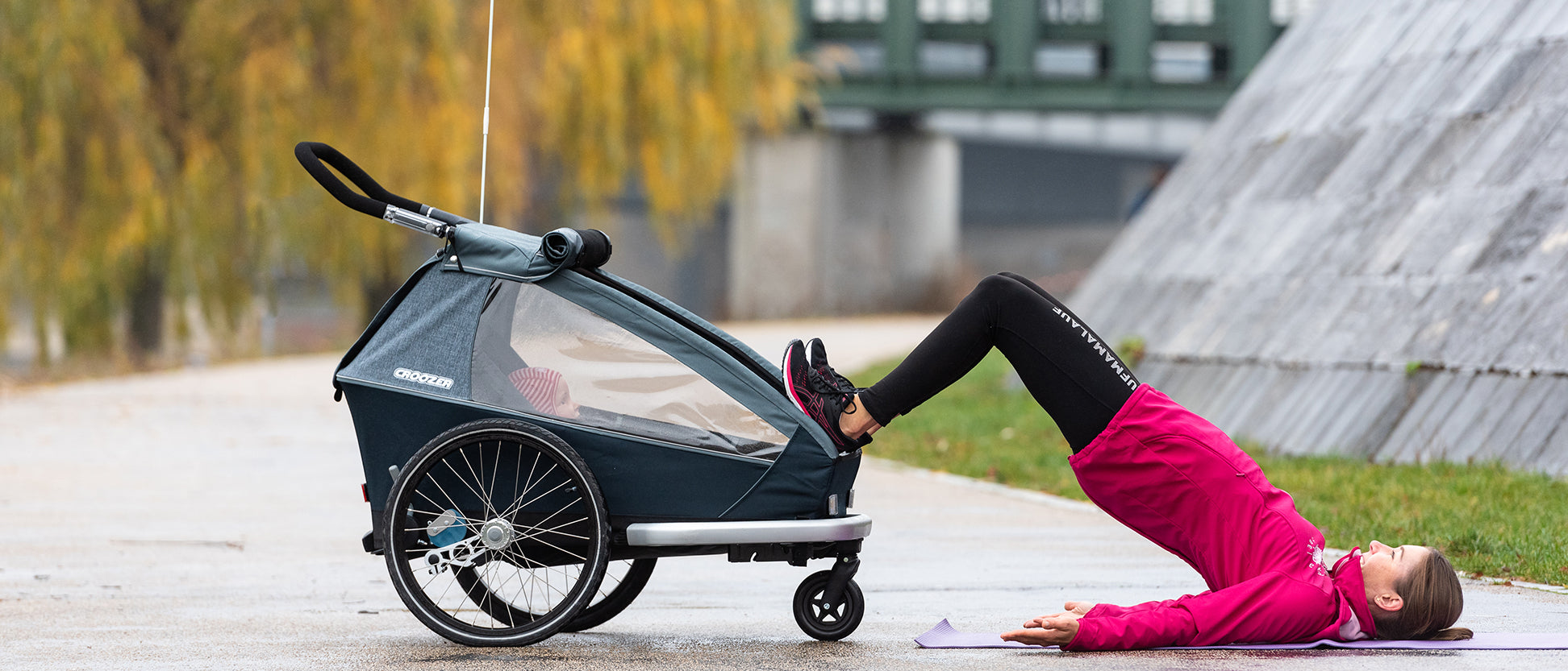 Woman performing leg raise exercise on yoga mat with feet on Croozer kid bike trailer in autumn setting